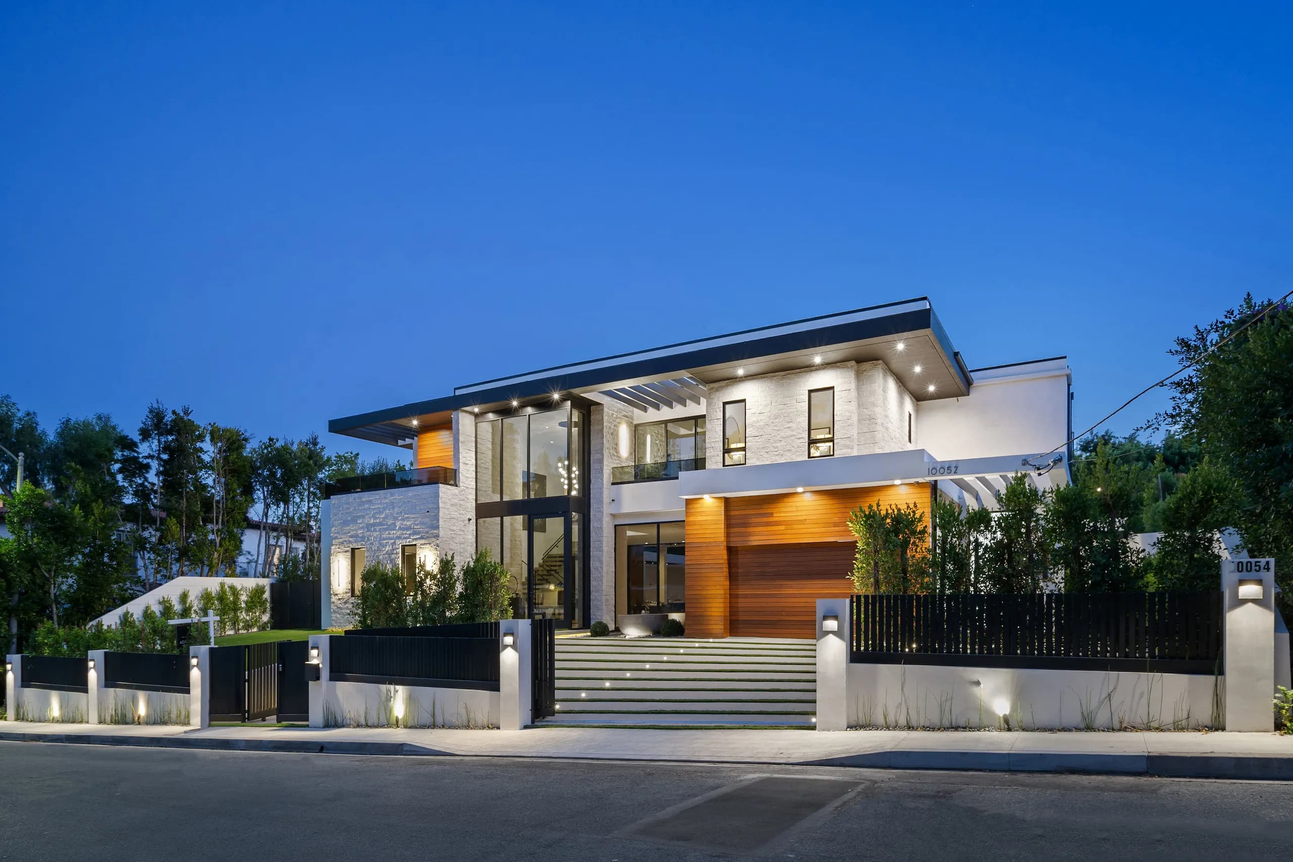 Contemporary luxury home with large glass entry, white stone walls, wood paneling, and front steps illuminated at dusk